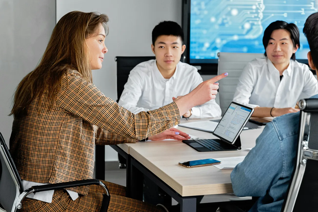 A woman in a plaid suit points while discussing with three colleagues at a table. Laptops and smartphones are visible, conveying a business setting.