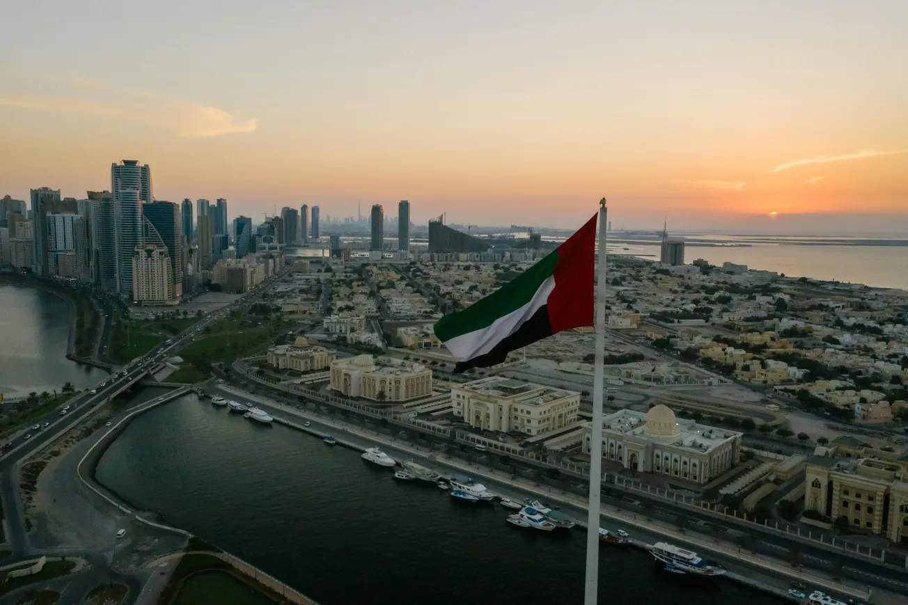 A UAE flag waves prominently over a cityscape at sunset, with high-rise buildings, a waterfront, and a warm, glowing sky, conveying a peaceful ambiance.