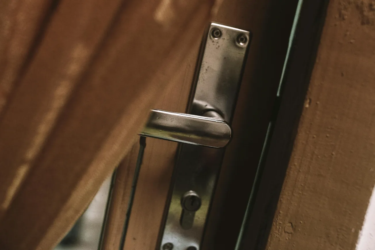 Close-up of a metallic door handle on a wooden door, slightly ajar. The image conveys a sense of anticipation and mystery.