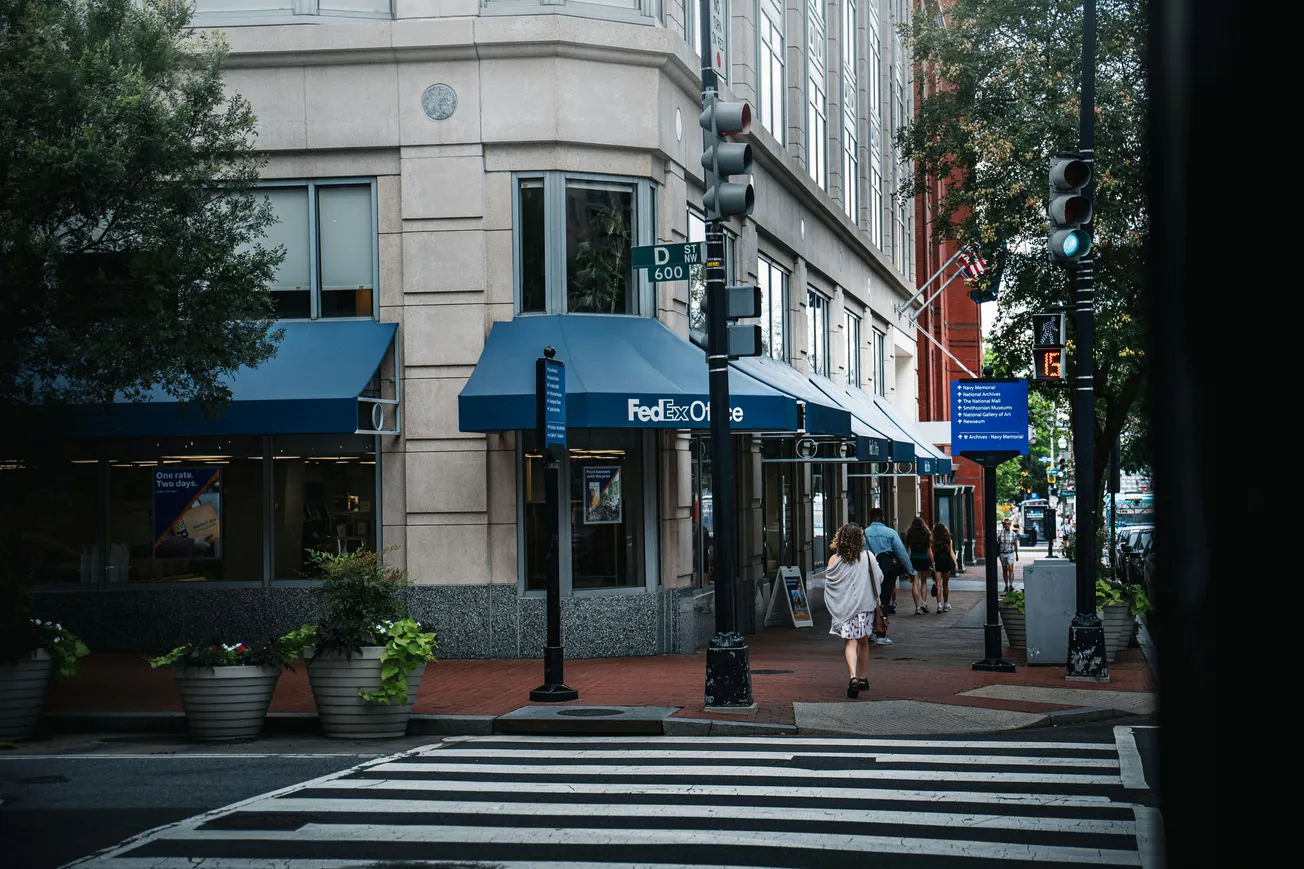 Street corner with a FedEx Office store featuring blue awnings on a cloudy day. A woman in a dress walks on the sidewalk, trees lining the path.