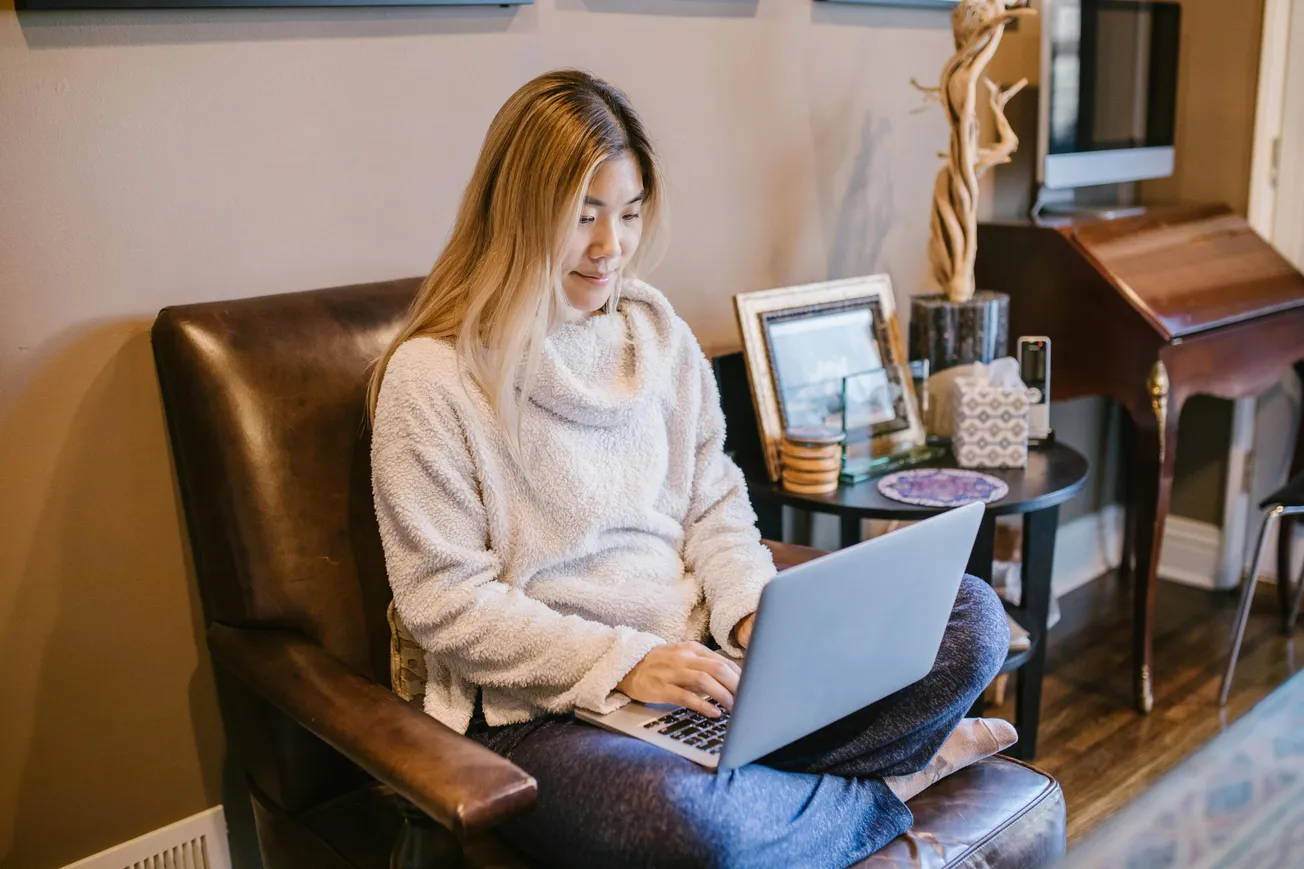 A woman in a cozy sweater sits cross-legged on a chair using a laptop. The room is warmly decorated with framed photos and a wooden sculpture on a side table.