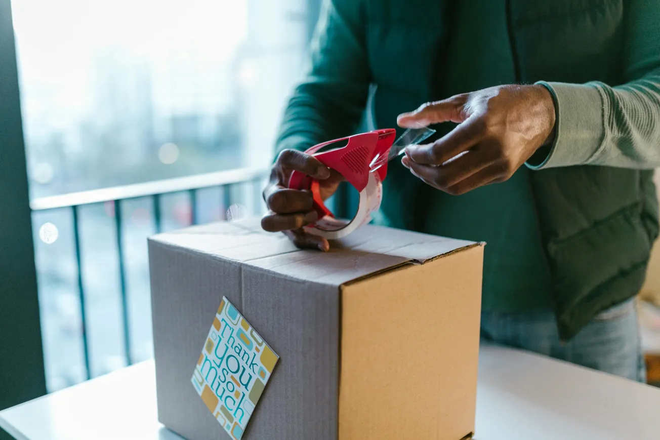 A person seals a brown cardboard box using a red tape dispenser. A card with "thank you so much" is attached. The setting is calm and bright.