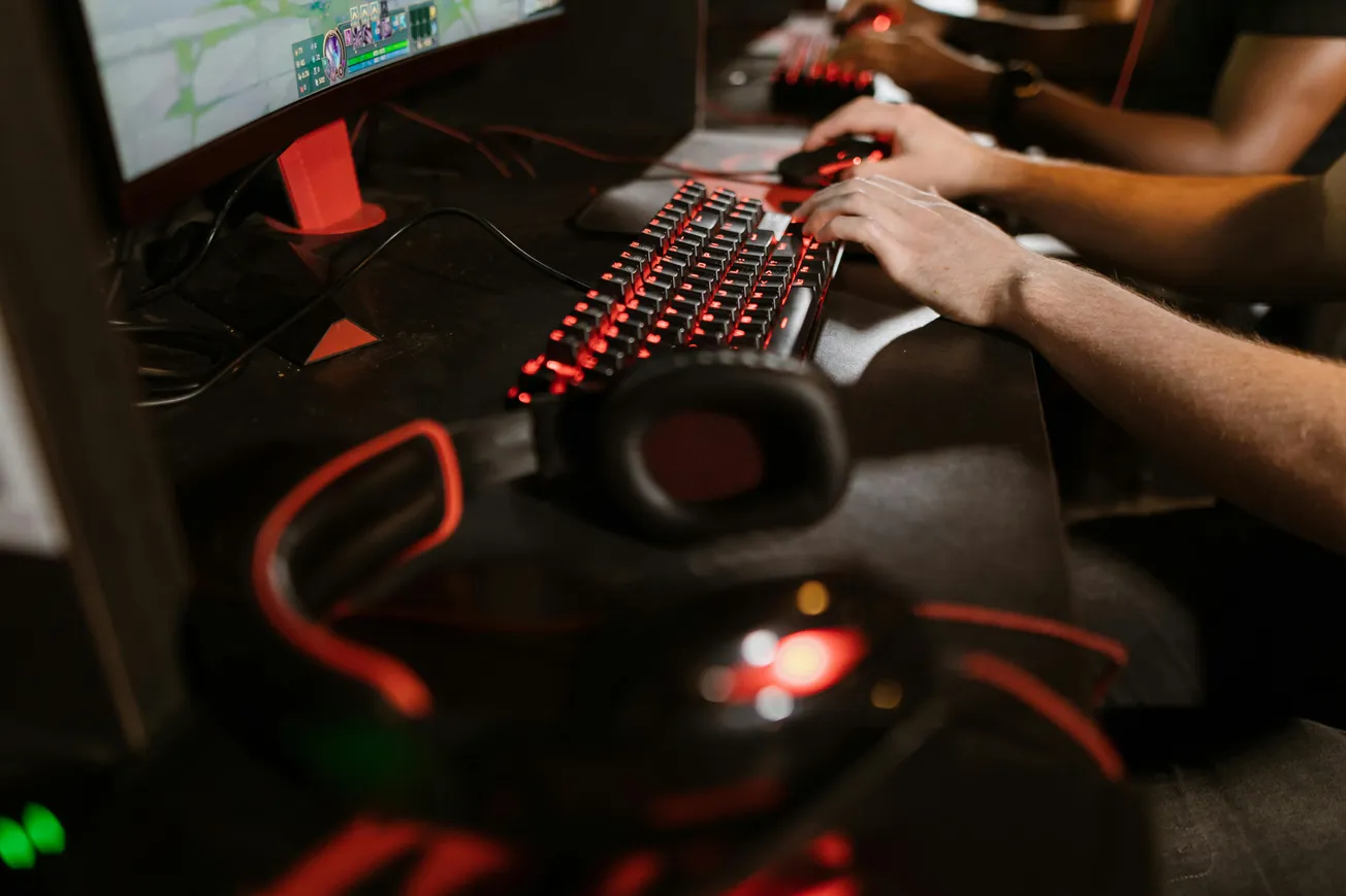 Close-up of gamers' hands using a computer with a glowing red keyboard and mouse. A large headset is visible, creating an intense gaming atmosphere.