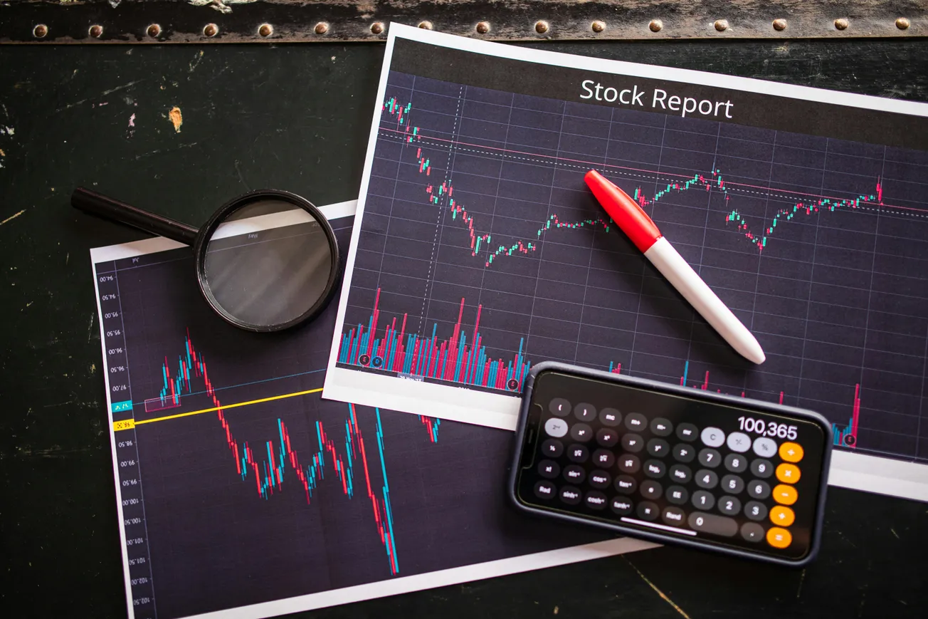 Stock report charts, a magnifying glass, pen, and smartphone calculator on a dark table. The mood is analytical, focused on financial data.