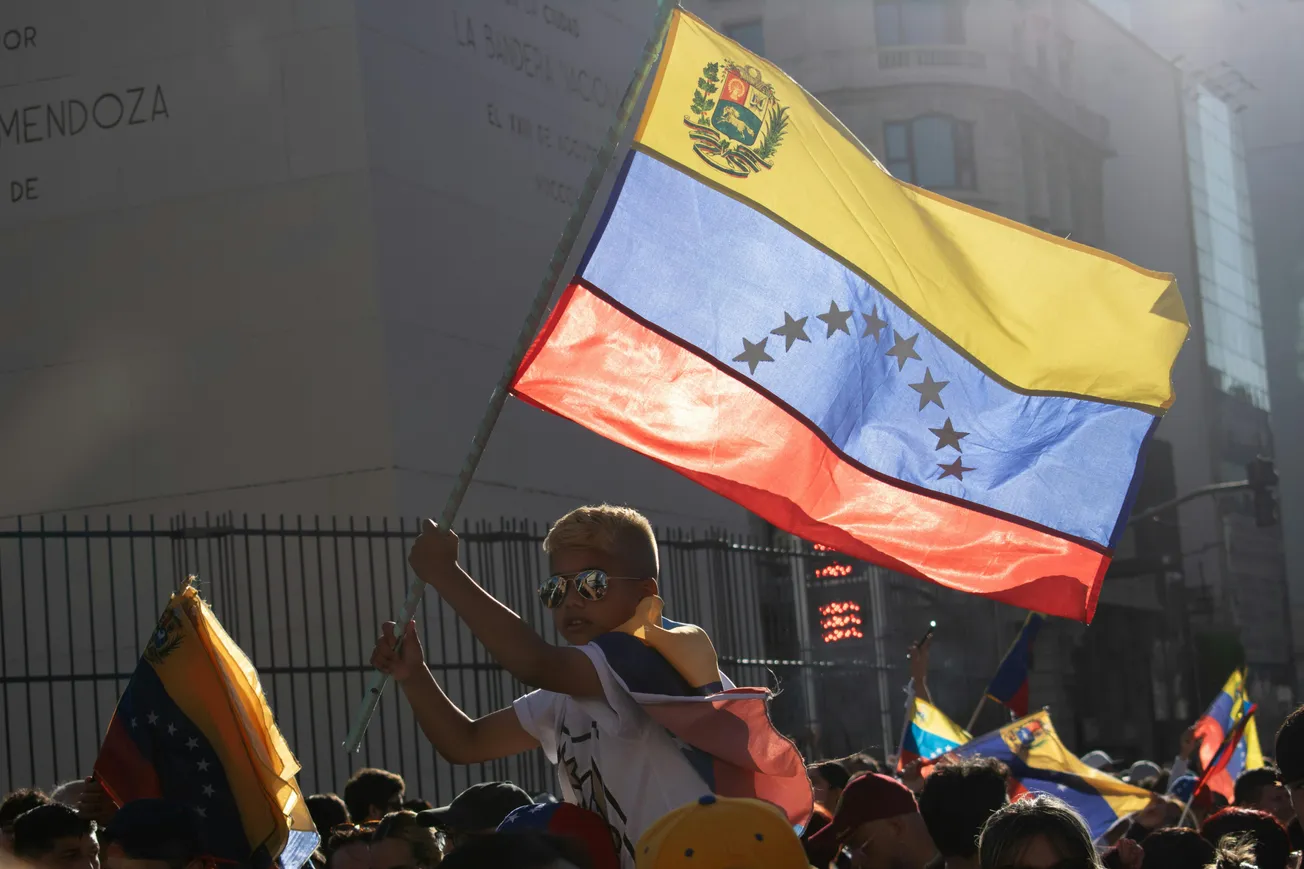 Boy wearing sunglasses holds a large Venezuelan flag at a lively outdoor rally. Bright sunlight enhances the energetic and hopeful atmosphere.