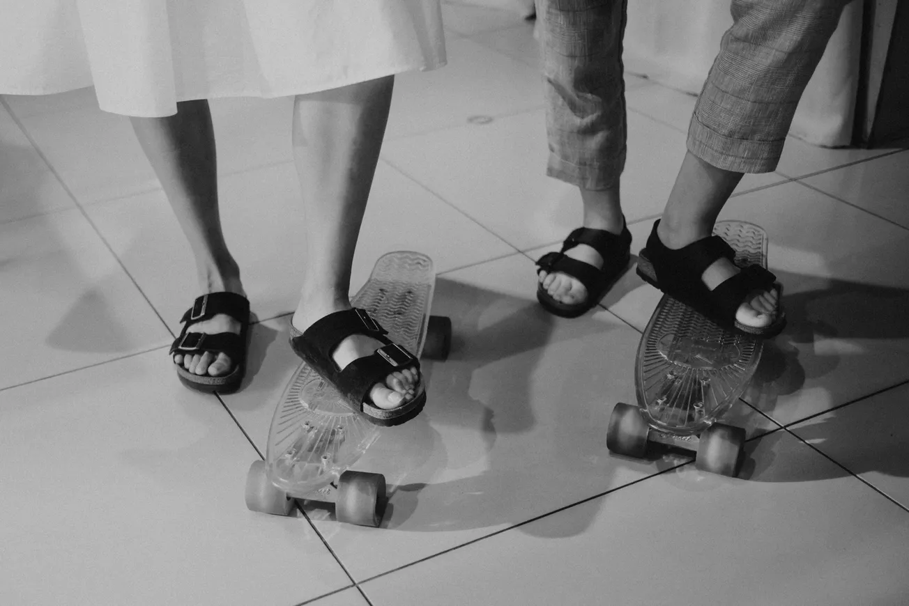 Two people stand with their feet on skateboards indoors. They're wearing sandals; one in a skirt, the other in pants. The tone is playful and casual.