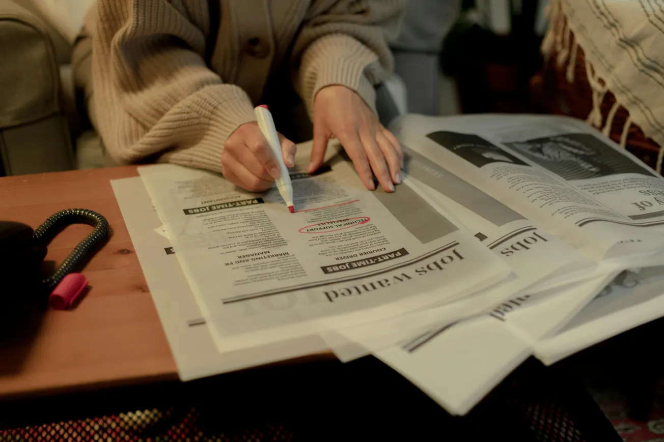 A person in a cozy sweater highlights job listings in a newspaper with a pink marker. Newspapers are spread across a wooden table, conveying focused determination.