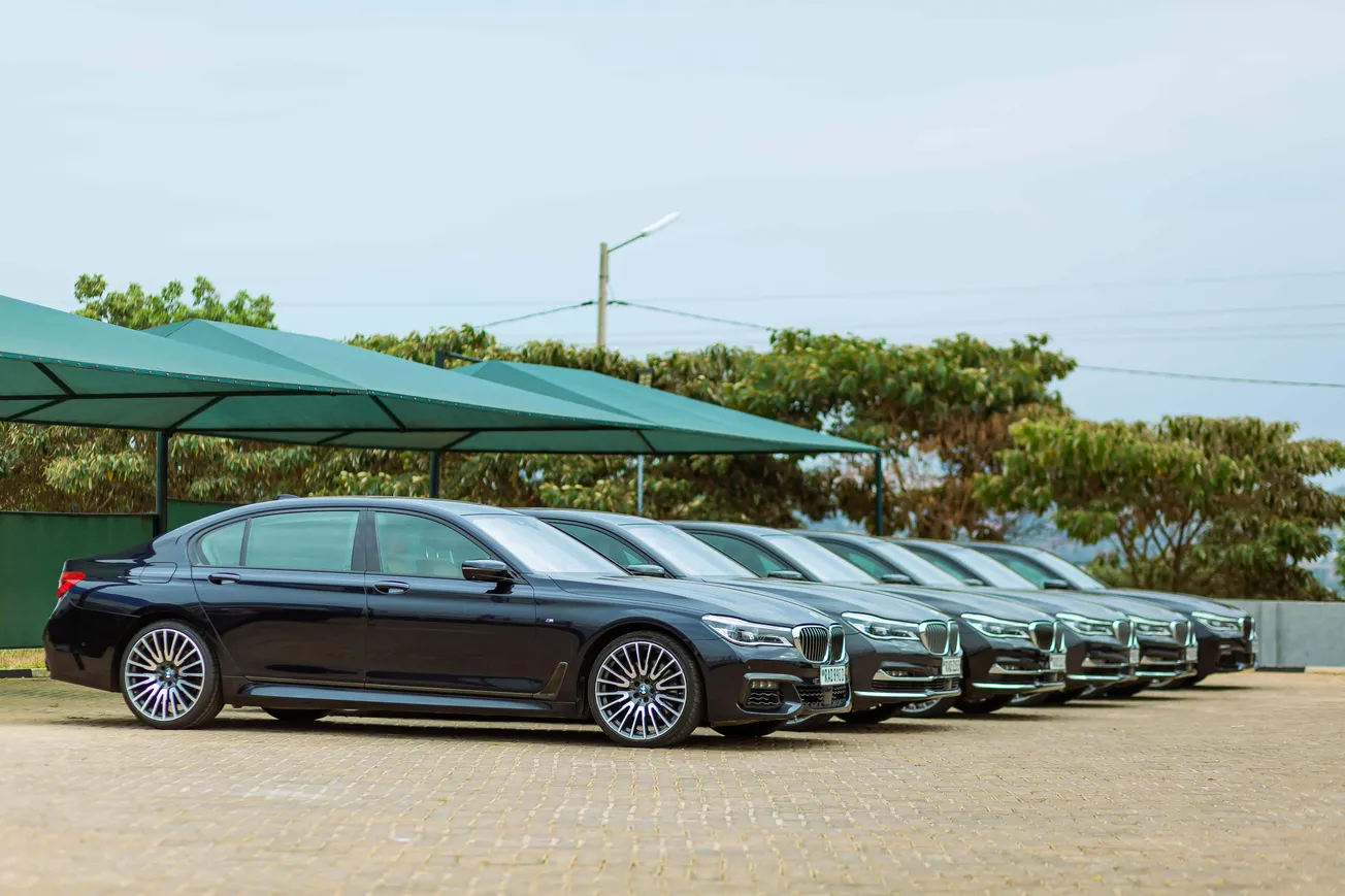 A line of six sleek black luxury sedans is parked under green canopies on a paved lot, with trees in the background under a clear sky.