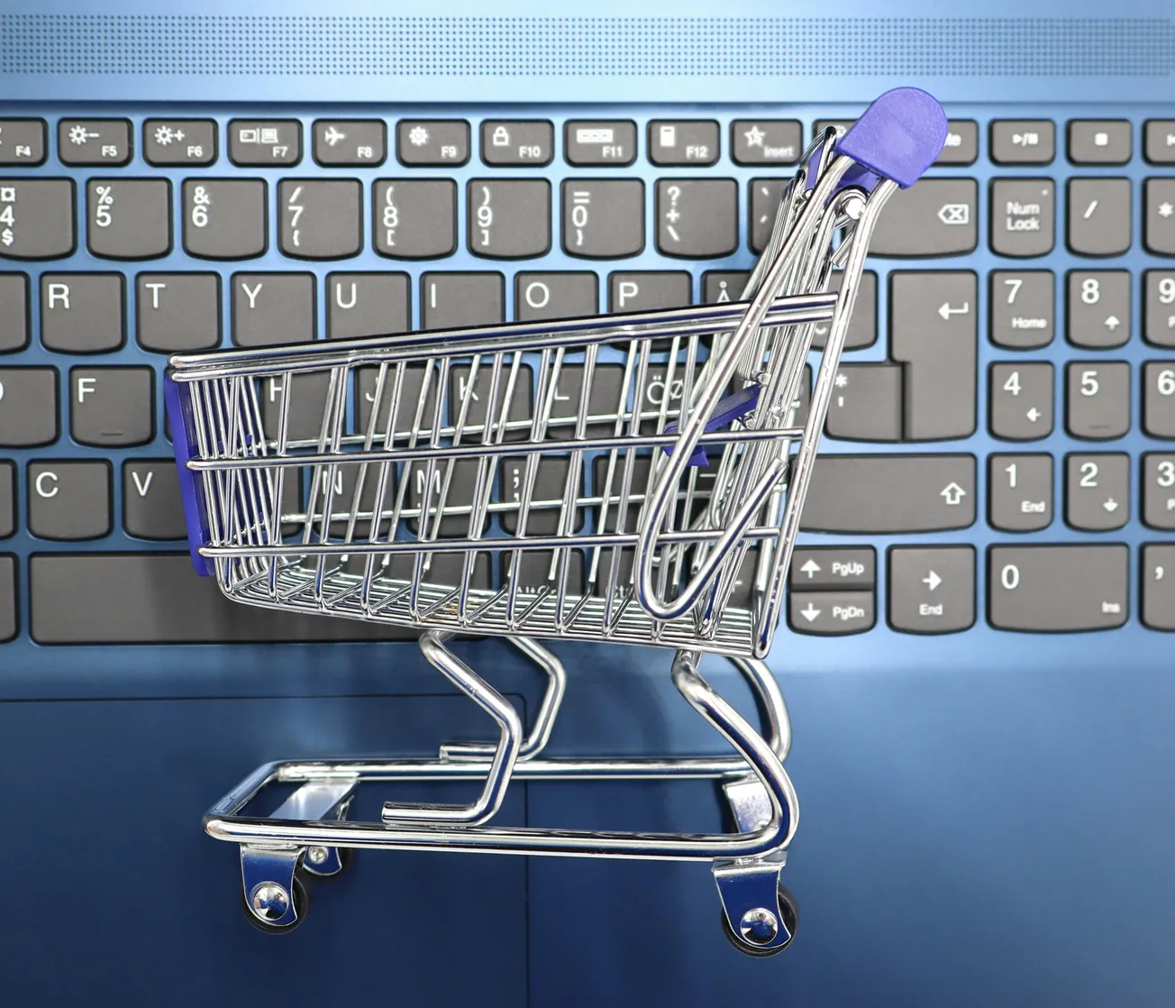 A small shopping cart sits on a blue laptop keyboard, symbolizing online shopping. The cart's metallic frame contrasts with the keyboard's gray keys.