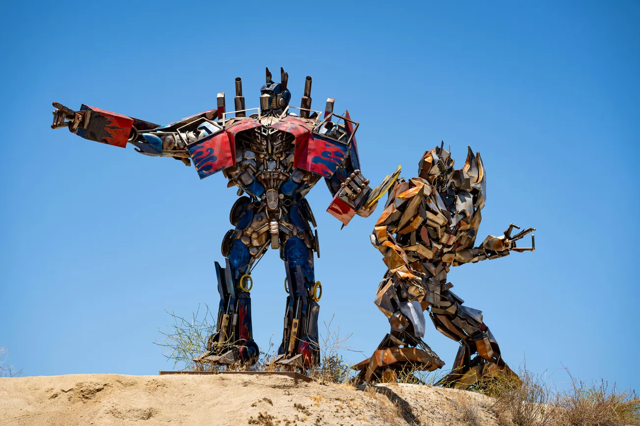 Two large, detailed robot sculptures stand on a sandy hill against a clear blue sky. One robot is red and blue; the other is silver and bronze.