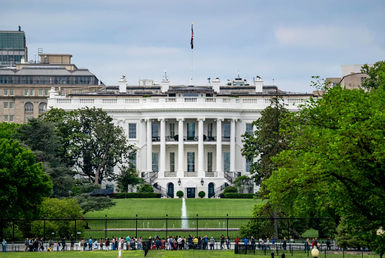 The image shows the White House, a large white building with columns, set against a backdrop of trees. A crowd is gathered in front, under a cloudy sky.