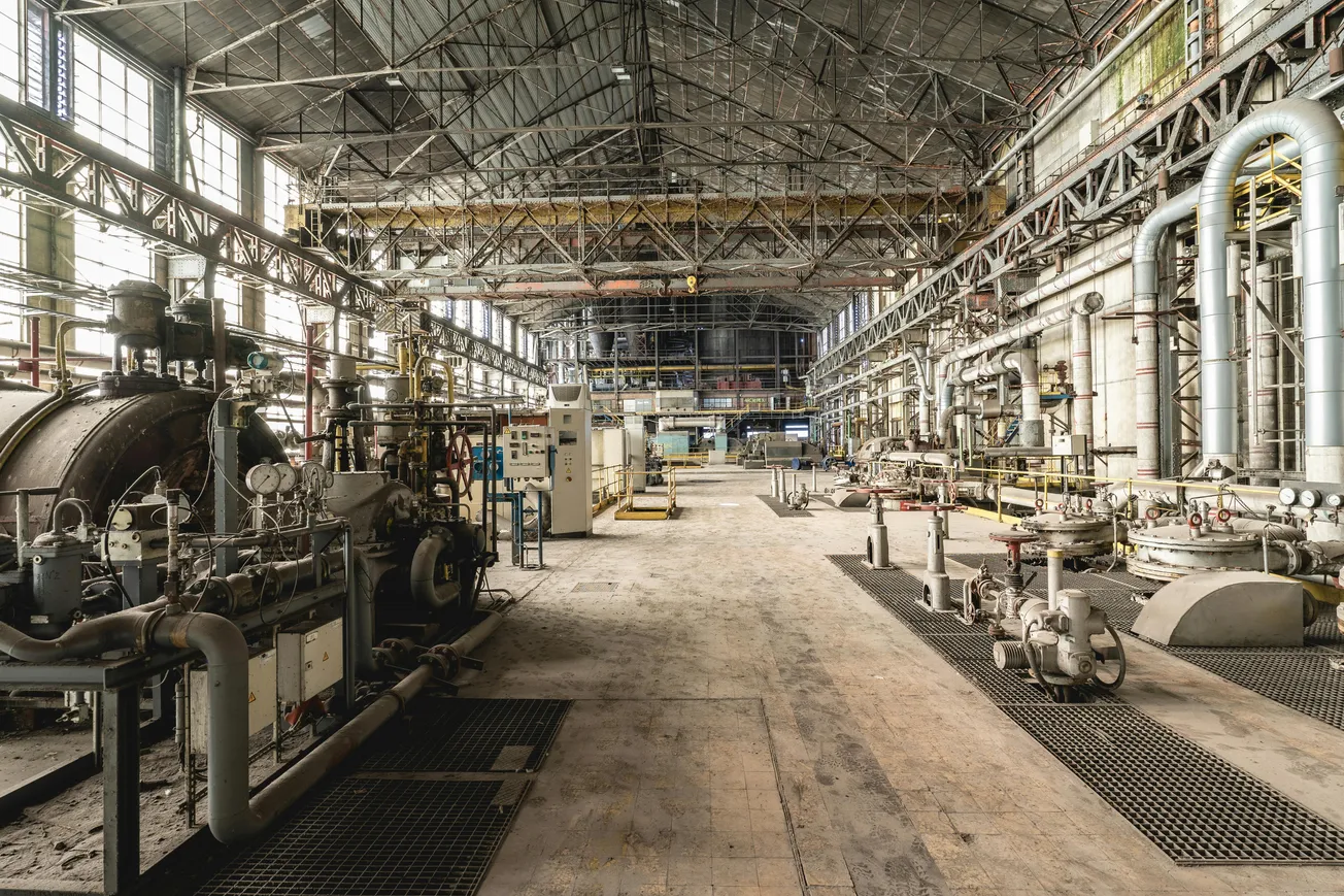 Spacious, industrial factory interior with large machinery, pipes, and metal girders. Dusty floors and subdued lighting create an abandoned feel.