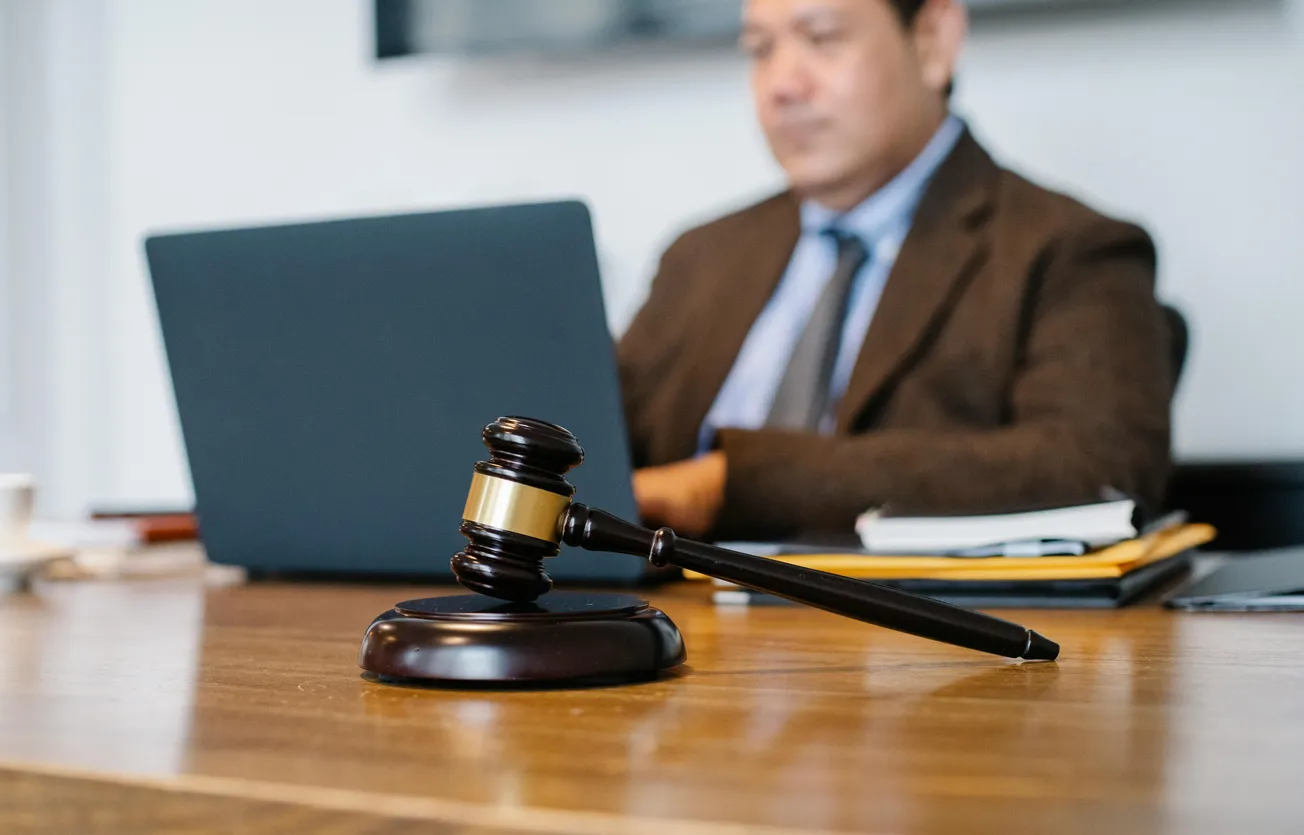 A wooden gavel rests on a table in focus, symbolizing justice. In the blurred background, a suited man works on a laptop, suggesting a legal setting.