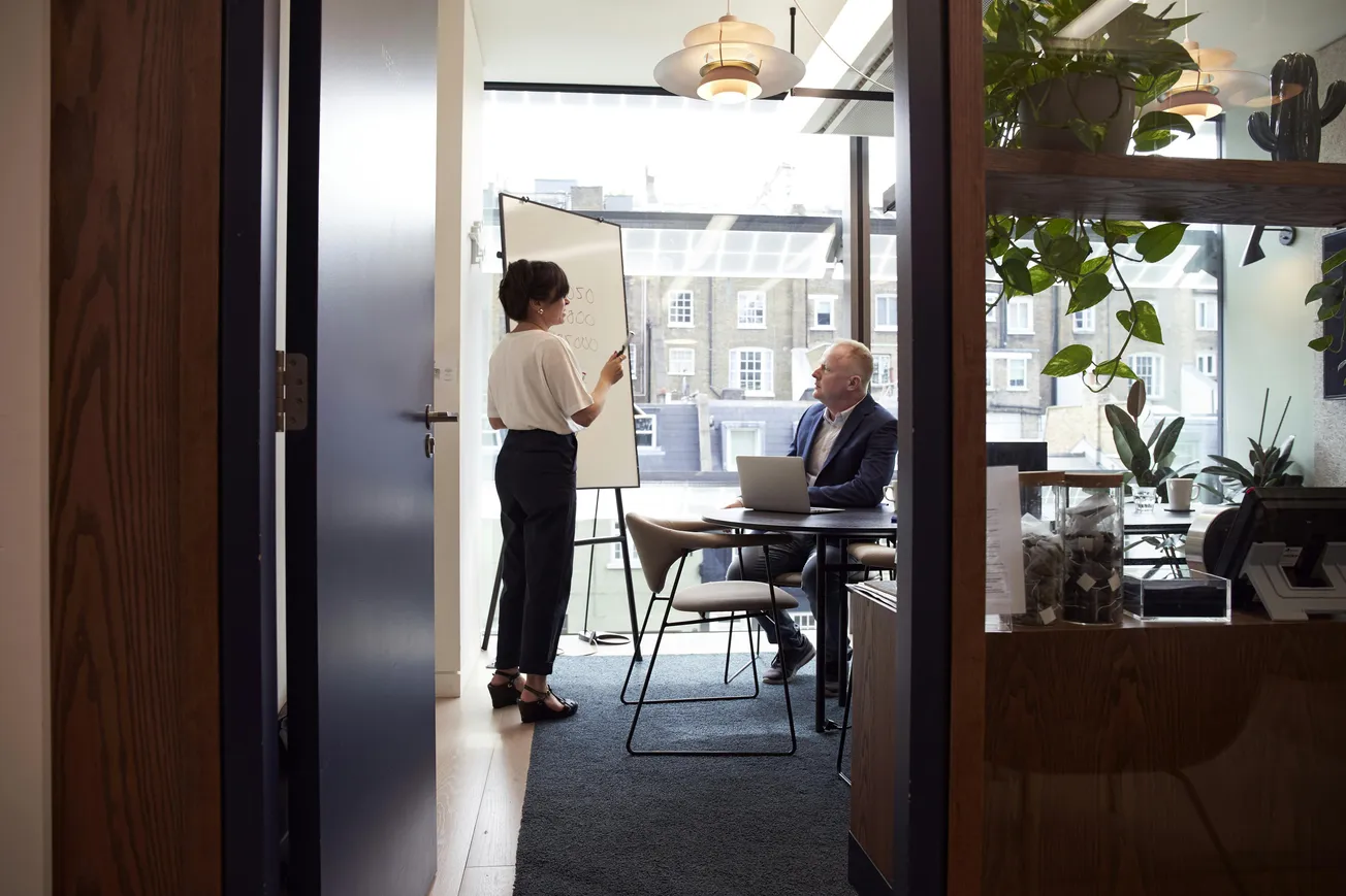 A woman presents at a whiteboard in a modern office, engaging with a seated man using a laptop. Large windows and plants create a bright, focused atmosphere.
