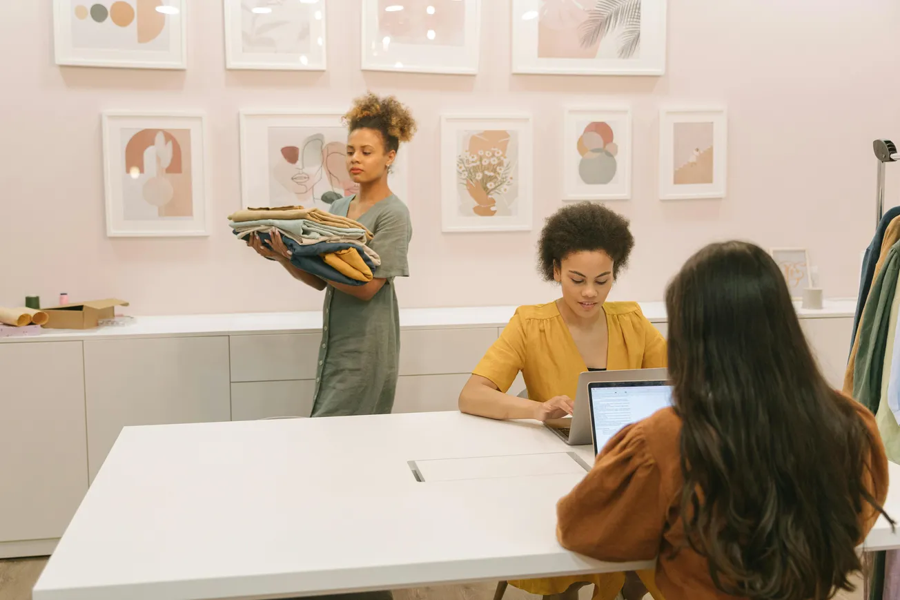 Three women work in a modern, minimalist room with pastel decor. One carries folded clothes, while two sit at a table with a laptop, focused and calm.
