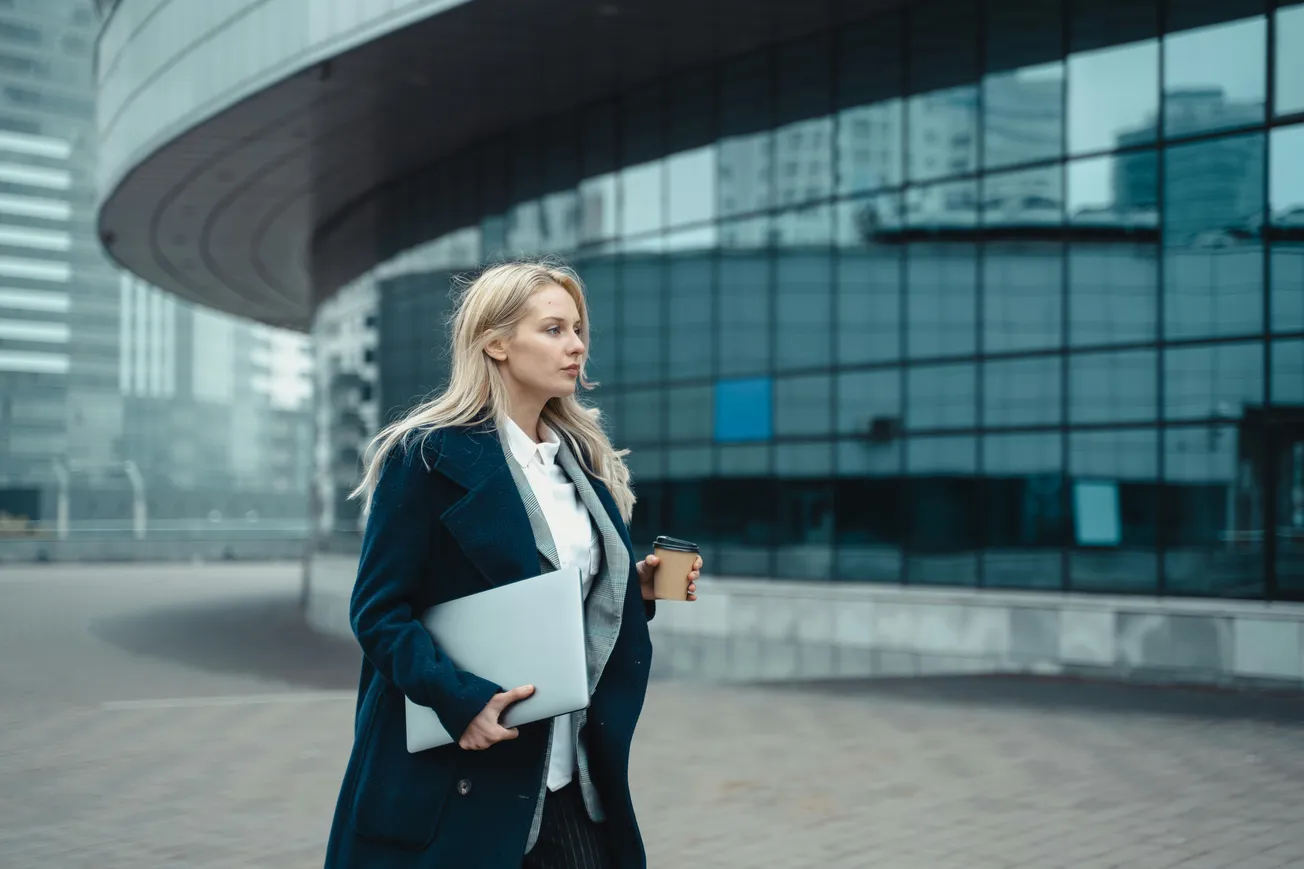 A woman in a blue coat walks outside a modern glass building, holding a laptop and a coffee cup. She appears focused and determined on a cloudy day.