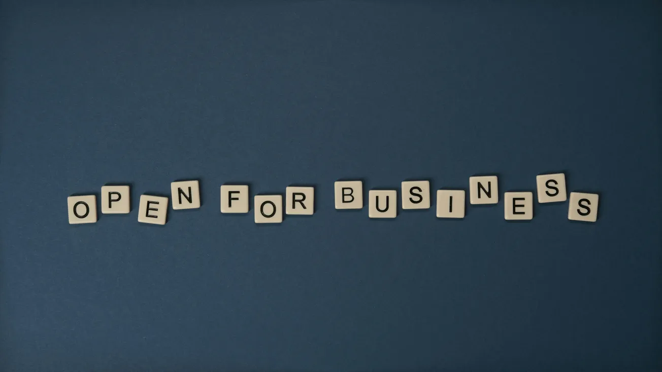 Scrabble tiles on a dark blue background spell "Open for Business." The arrangement conveys readiness and welcome, suggesting a positive, inviting tone.