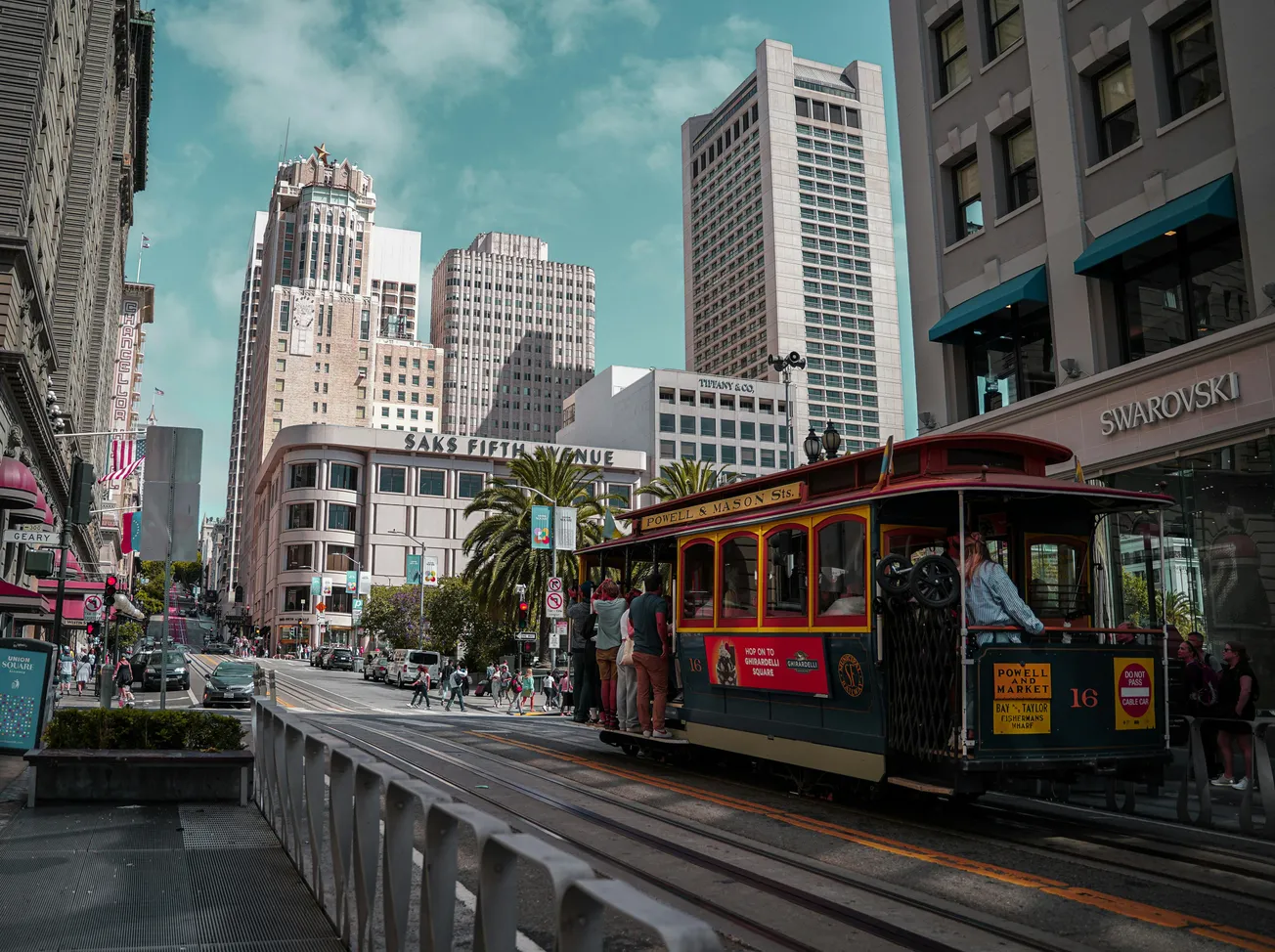 Street scene in a bustling city with a classic red cable car and people riding it. Tall buildings under a clear sky create an urban vibe.