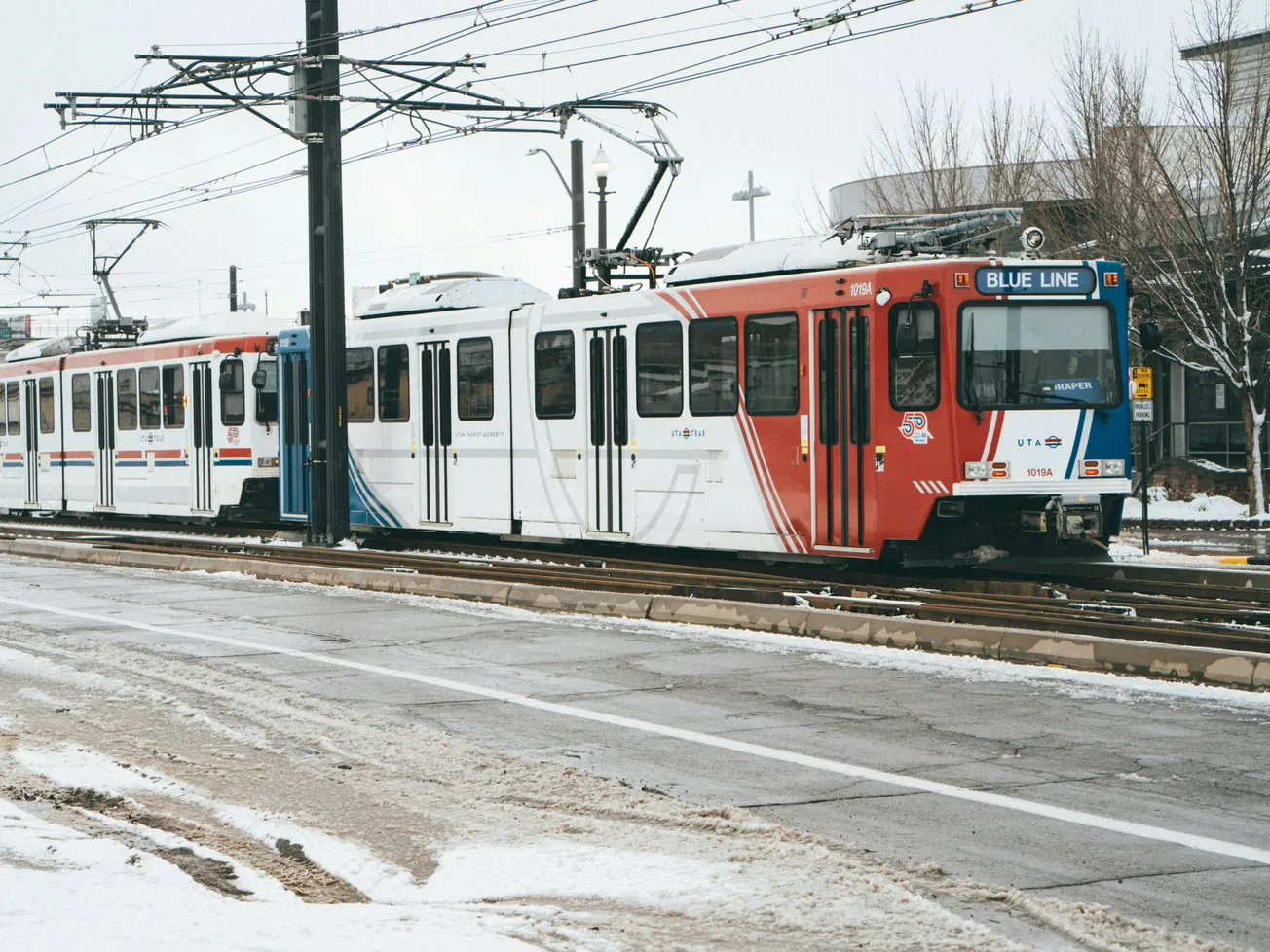 A light rail train on the snowy Blue Line track stops at a station. The sky is overcast, and bare trees surround the area, creating a wintery atmosphere.
