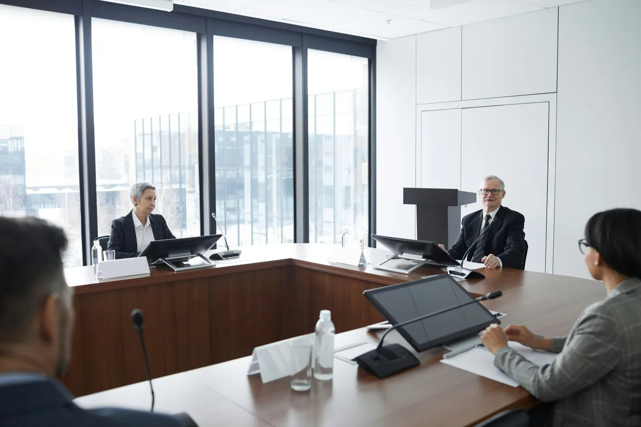 Four people in a modern conference room with large windows are engaged in a meeting. The tone is professional and collaborative.