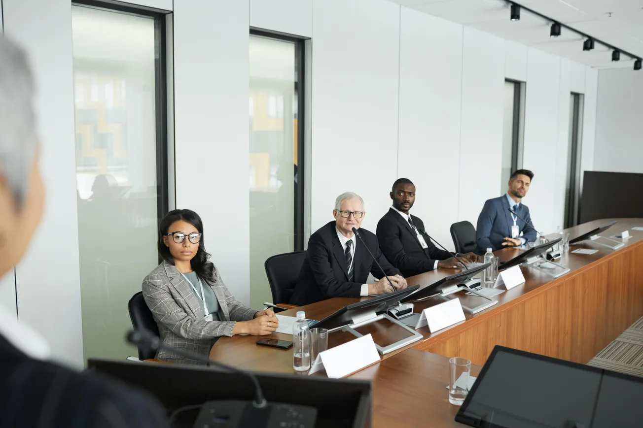 A diverse group of four professionals, seated at a long conference table, attentively listen to a speaker. The setting suggests a formal meeting.