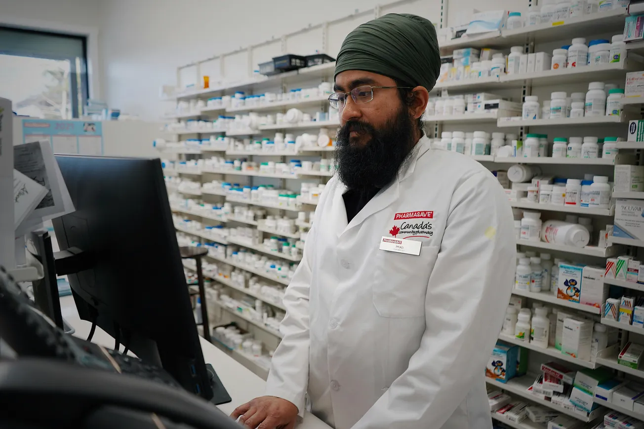 A pharmacist in a white coat, wearing a turban, stands at a computer in a pharmacy. Shelves filled with medicine bottles line the background. The atmosphere is professional and focused.
