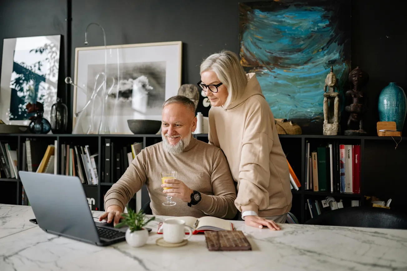 A man and woman smile while using a laptop in a cozy room. The man holds a wine glass; shelves with art and books fill the background, creating a warm ambiance.