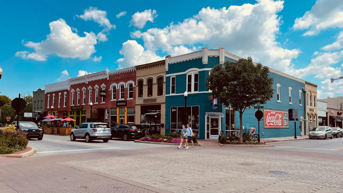 A vibrant street corner features colorful historic buildings under a bright blue sky with fluffy clouds. People, cars, and a Coca-Cola mural add life and nostalgia.