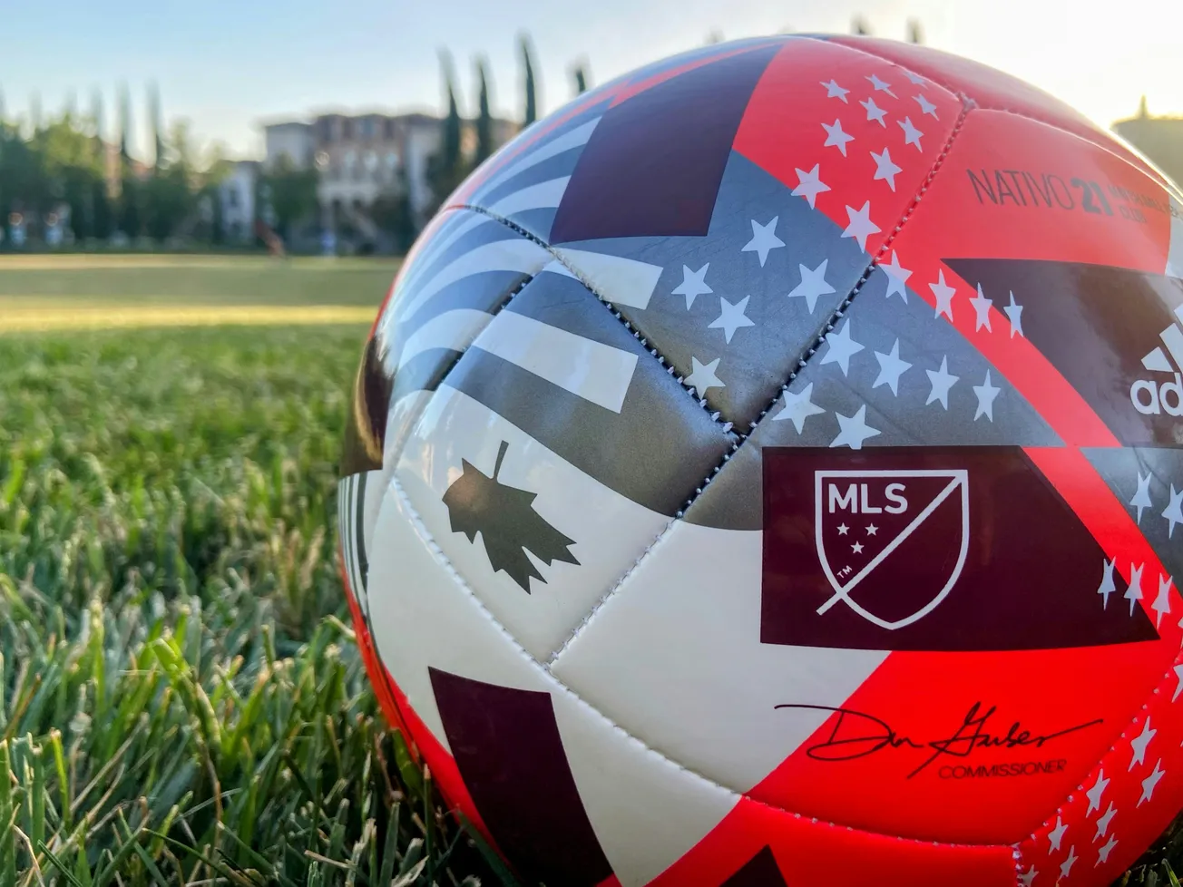 Close-up of a colorful soccer ball featuring the MLS logo and American/Canadian flag design, resting on green grass with blurred trees and buildings in the background.