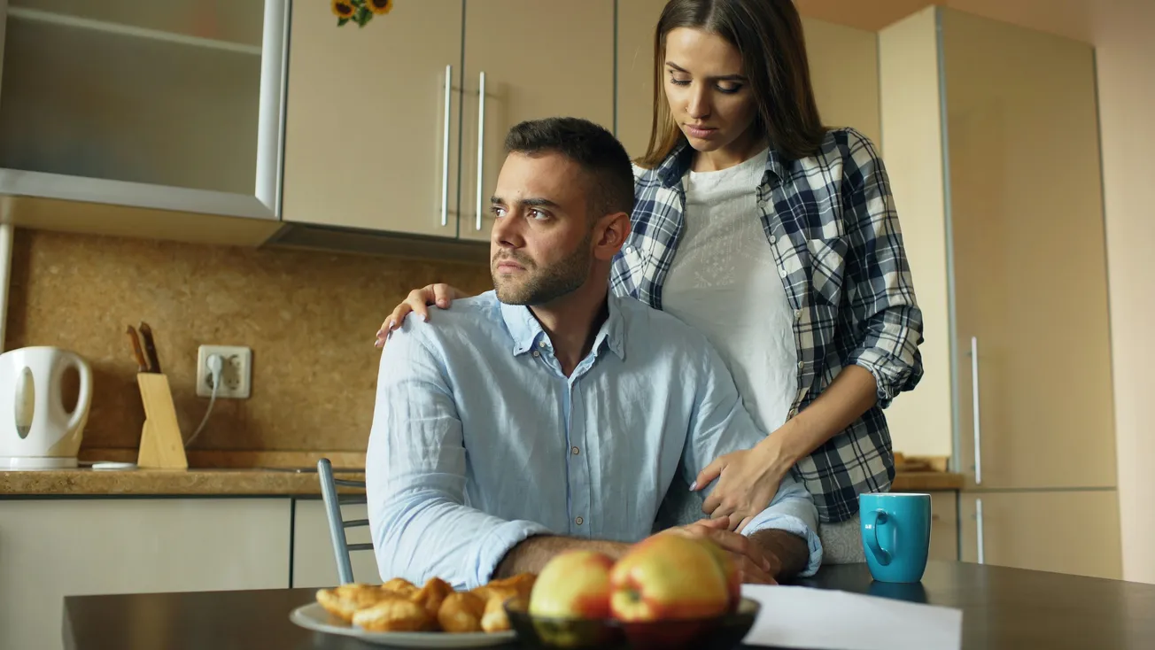 A concerned woman comforts a seated man in a kitchen, her hand on his shoulder. A plate of pastries and apples sits on the table, next to a blue mug.