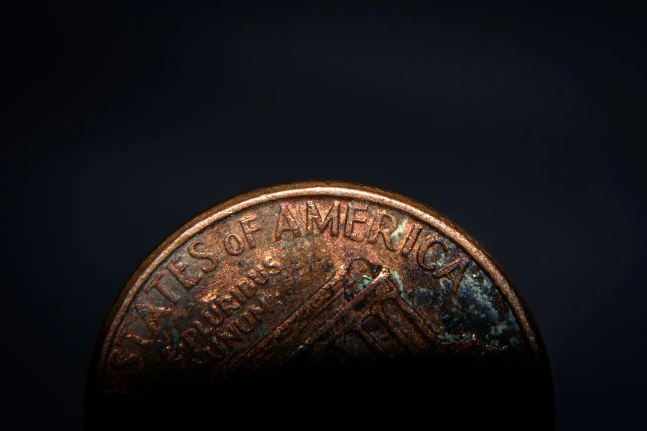 Close-up of a partially-lit, weathered US penny, with the top half visible, showing the words "States of America" against a dark background.