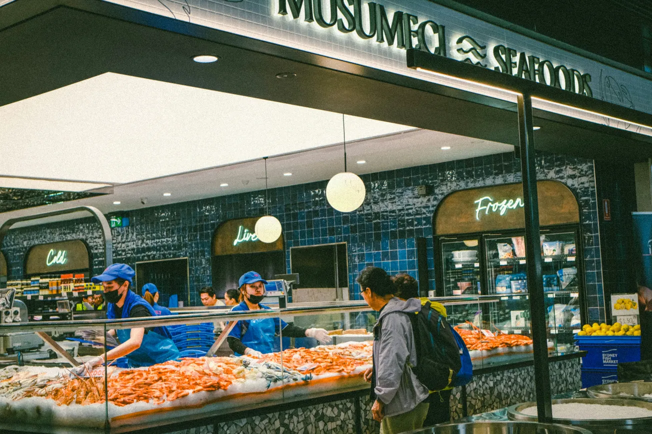 A bustling seafood market with workers in blue uniforms assisting customers. Iced displays showcase fresh fish and shrimp. The atmosphere feels busy and lively.