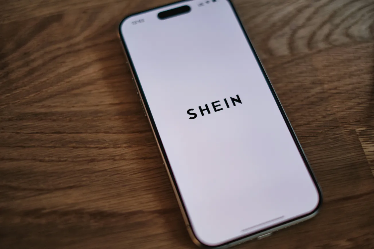 Smartphone on a wooden table showing "SHEIN" on its white screen, suggesting an online shopping app. The scene conveys modern, digital retail.