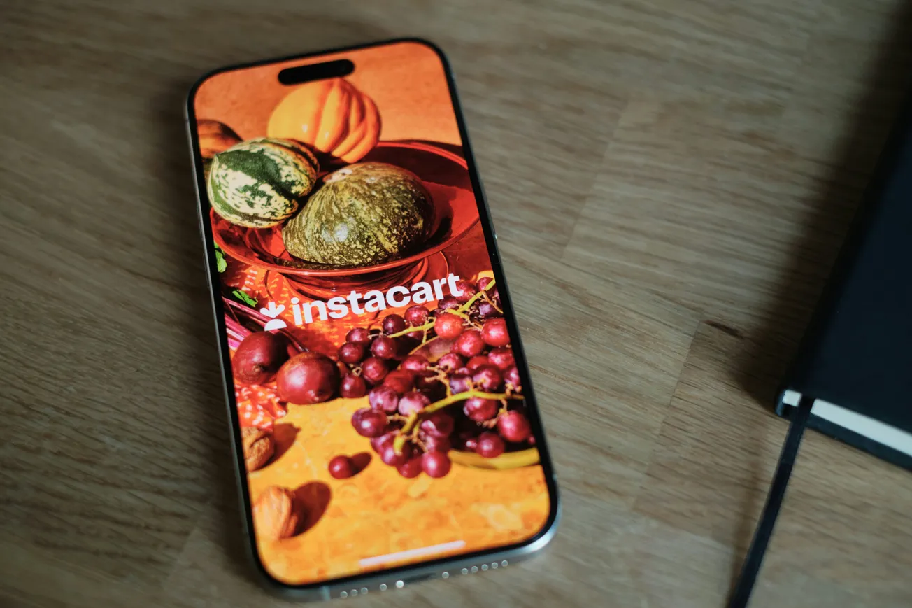 A smartphone on a wooden table displays the Instacart app with an image of assorted fruits and vegetables, conveying a fresh, vibrant feel.
