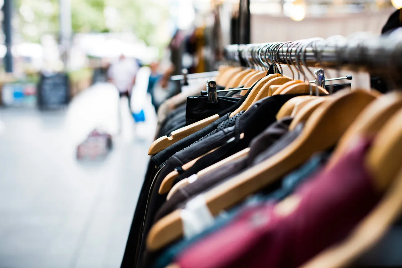 Close-up of a clothing rack with various garments on hangers in a fashion store. The background is blurred, suggesting a busy shopping environment.
