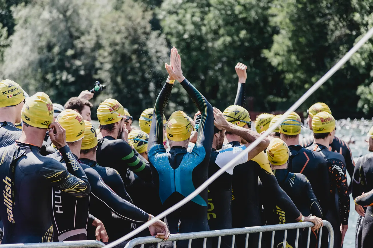 A group of athletes in wetsuits and yellow swim caps gather near a metal railing, preparing for a swim race. The mood is focused and energetic.