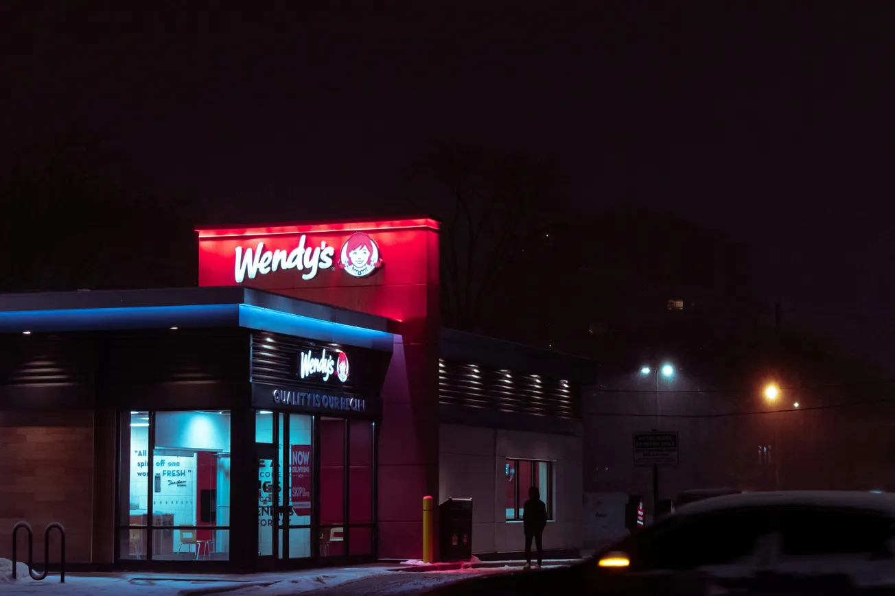 A Wendy's restaurant is lit up at night, with a glowing red sign against a dark, snowy backdrop. Few cars and a silhouette of a person create a quiet, moody atmosphere.