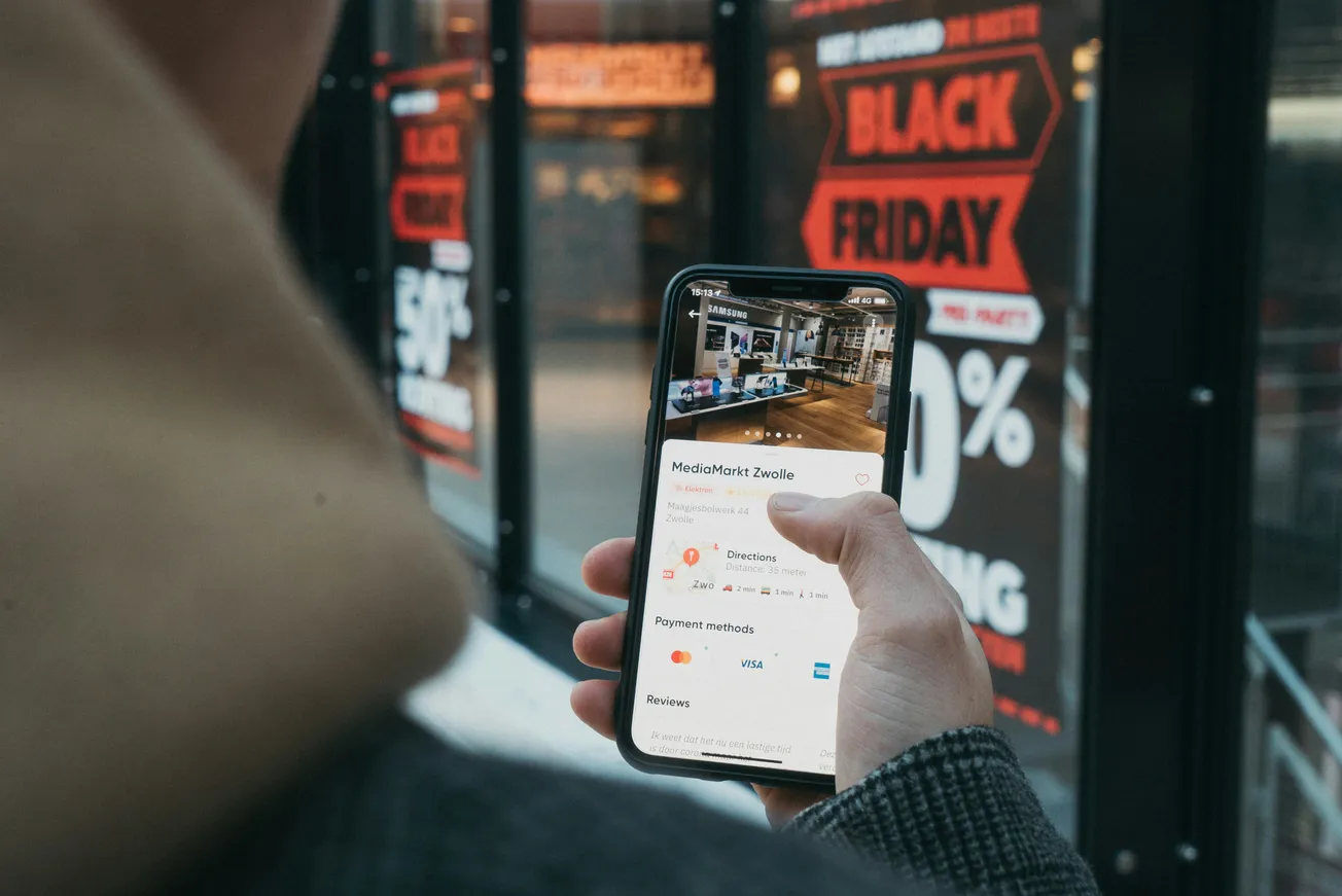 A person holds a smartphone showing store directions, standing in front of a shop window with prominent "Black Friday" sale signs, creating a shopping vibe.