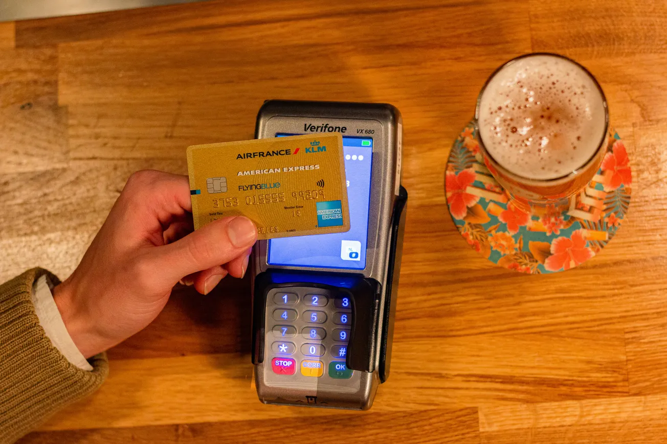 A person holds a yellow credit card near a payment terminal on a wooden table. Beside it, a frothy drink sits on a colorful floral coaster.