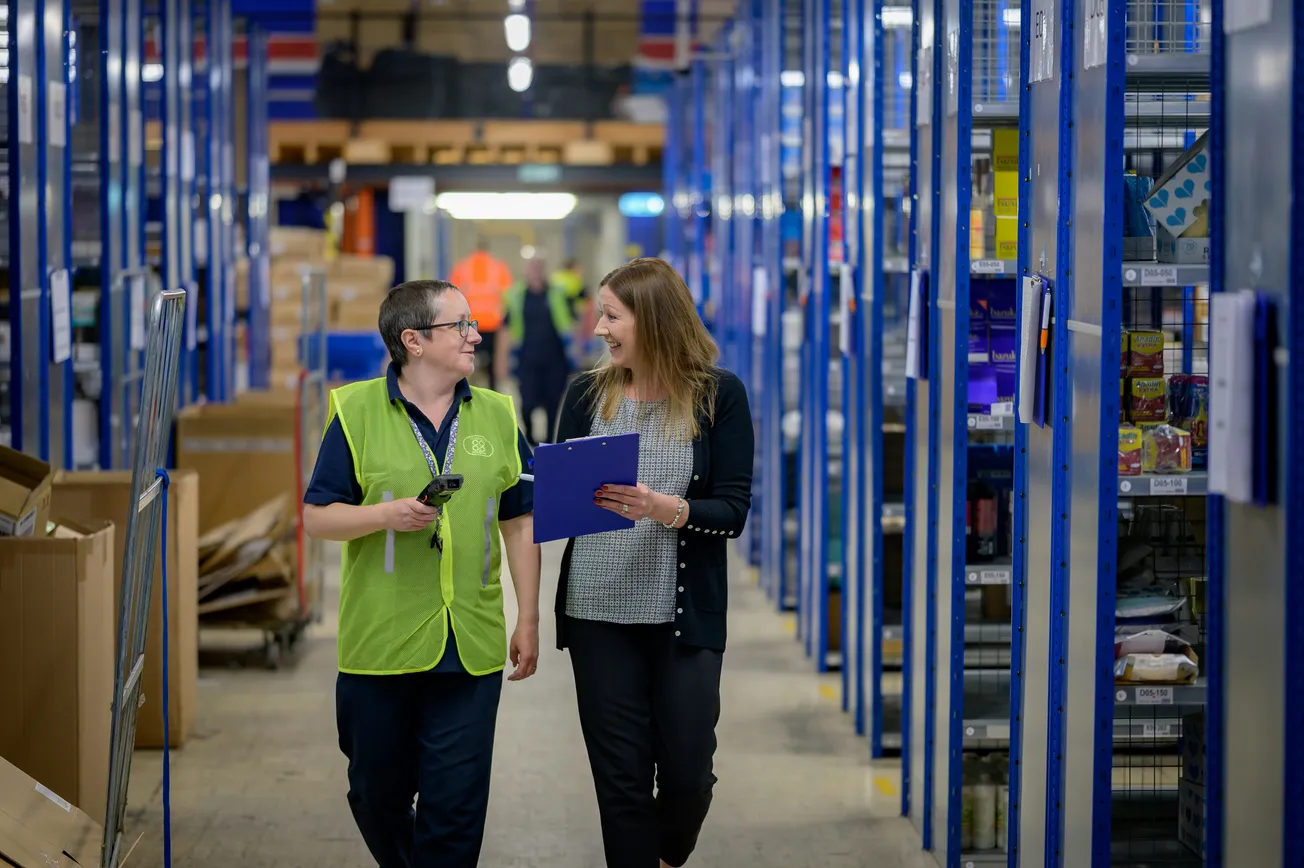 Two people walk through a warehouse aisle, engaging in conversation. One wears a neon vest, holding a device, while the other carries a clipboard, smiling.