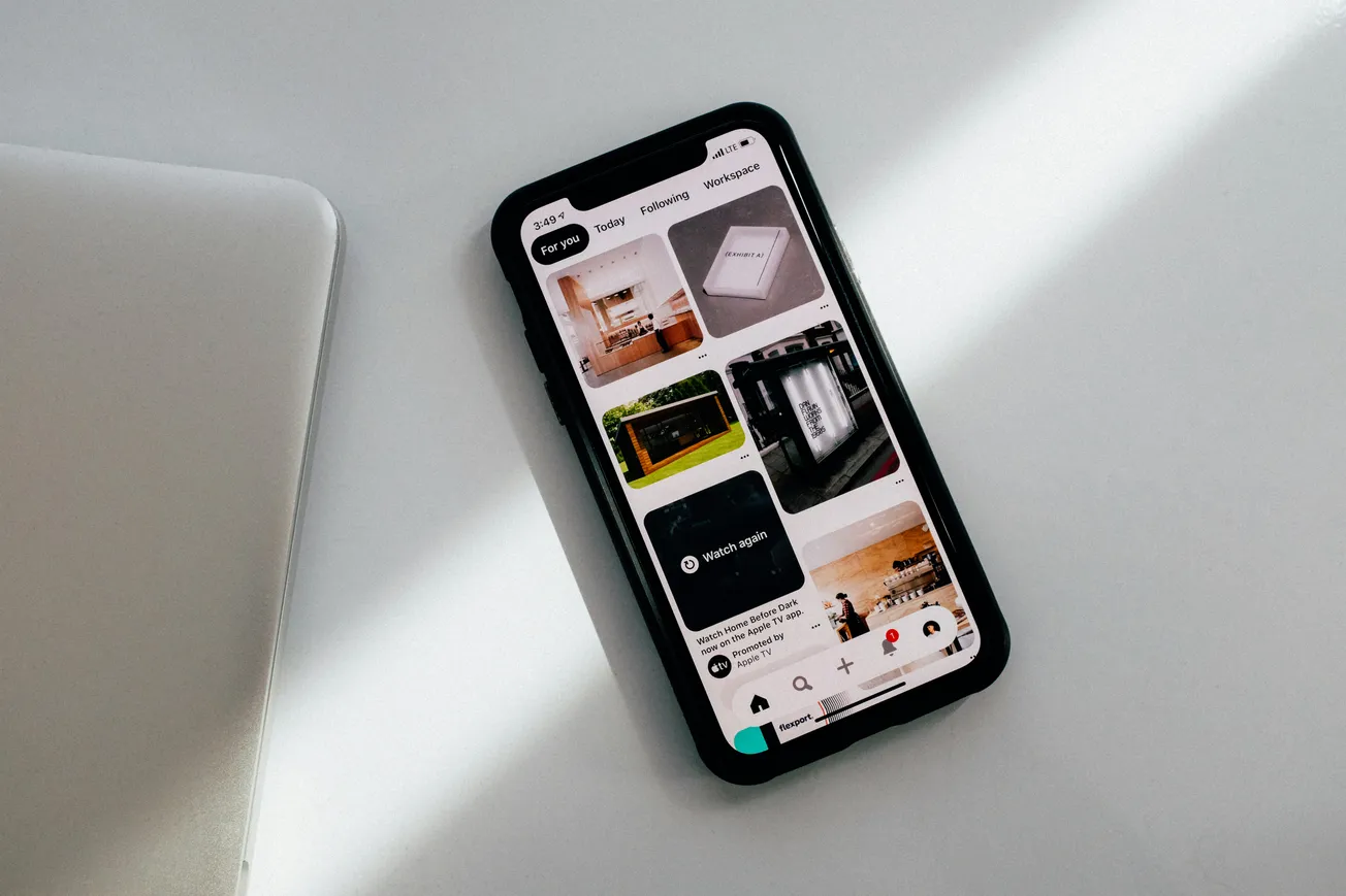 Smartphone on a white surface displays a Pinterest feed with home decor images. Soft light casts shadows, creating a calm, minimalistic atmosphere.