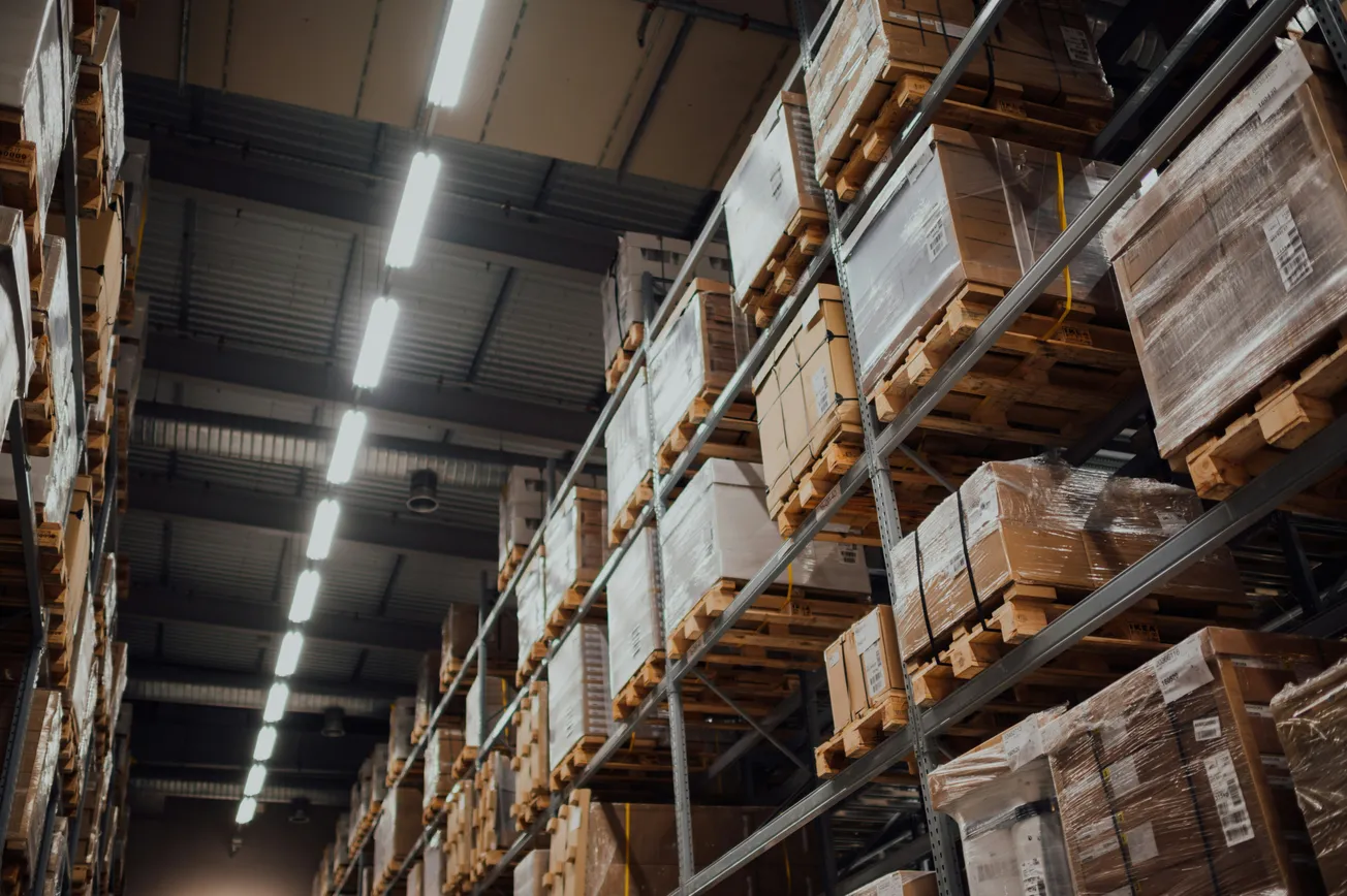 Warehouse interior with tall shelves filled with stacked, wrapped cardboard boxes on wooden pallets. Fluorescent lights illuminate the spacious area.