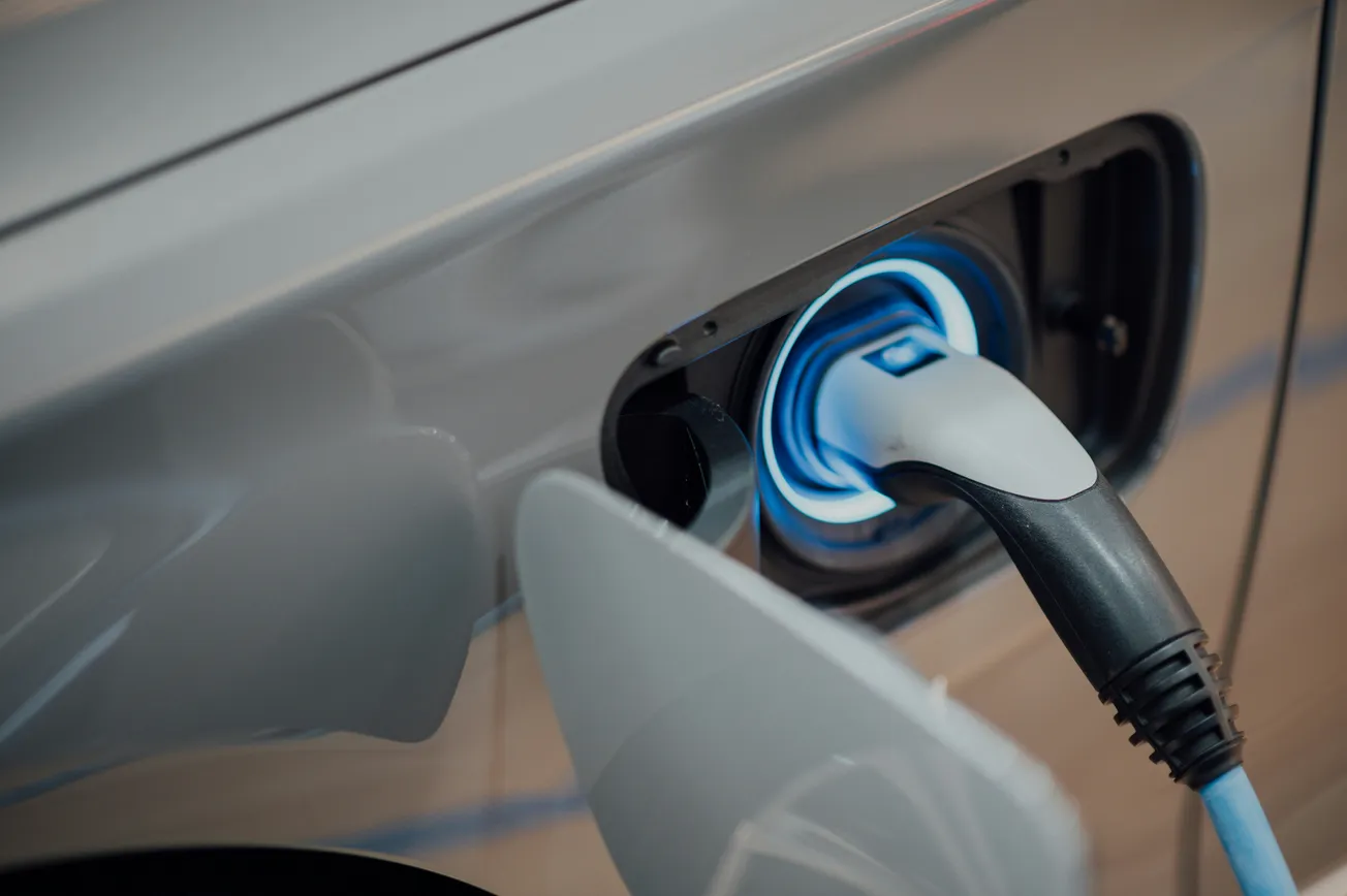 Close-up of an electric vehicle charging port with a cable plugged in, glowing blue. The sleek car surface indicates modern technology and sustainability.