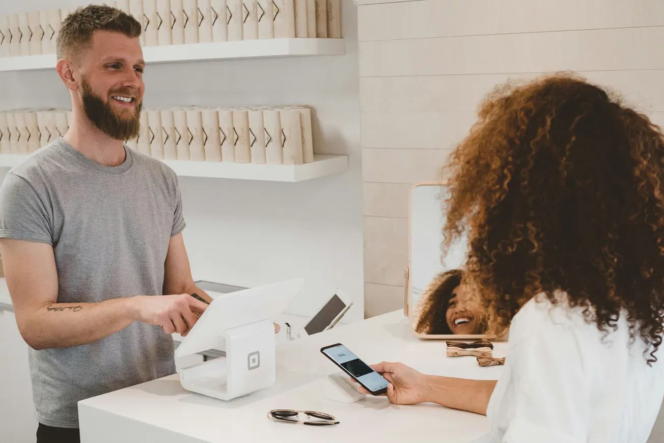 A smiling cashier with a beard stands behind a counter using a sleek white tablet-based register. A customer with curly hair pays using a smartphone. The setting is bright and modern.