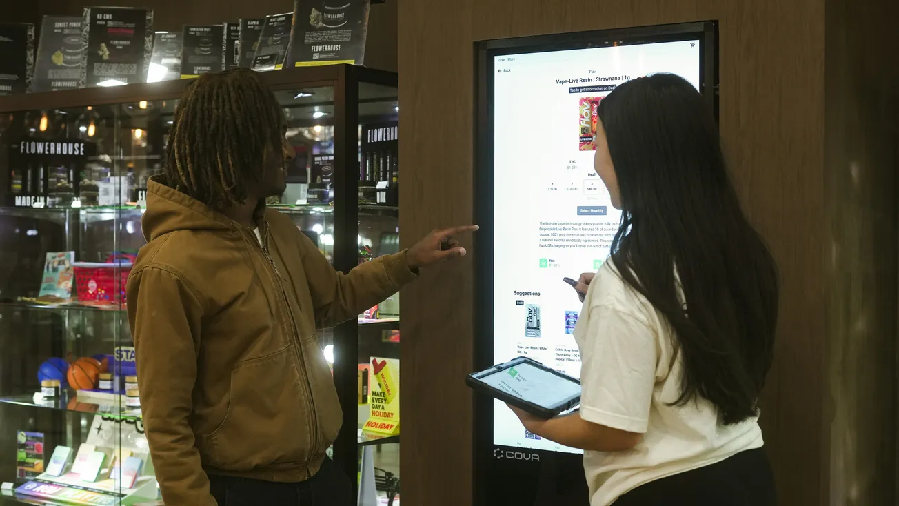 Two people stand in front of a digital kiosk inside a store. One points at the screen, while the other holds a tablet, discussing product details.