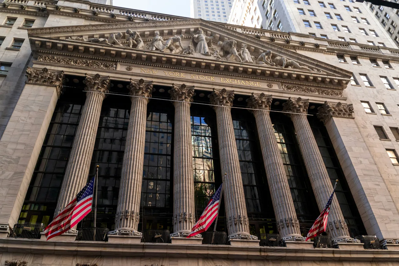 Facade of the New York Stock Exchange with six grand columns and statues above. Three American flags fly in front, conveying formality and prestige.
