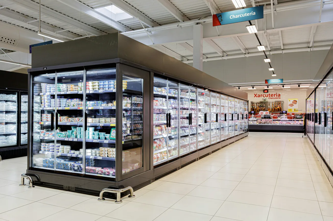 Grocery store aisle with glass-door freezers densely stocked with various products. "Charcutería" sign hangs above, leading to a meat counter. Bright, clean setting.