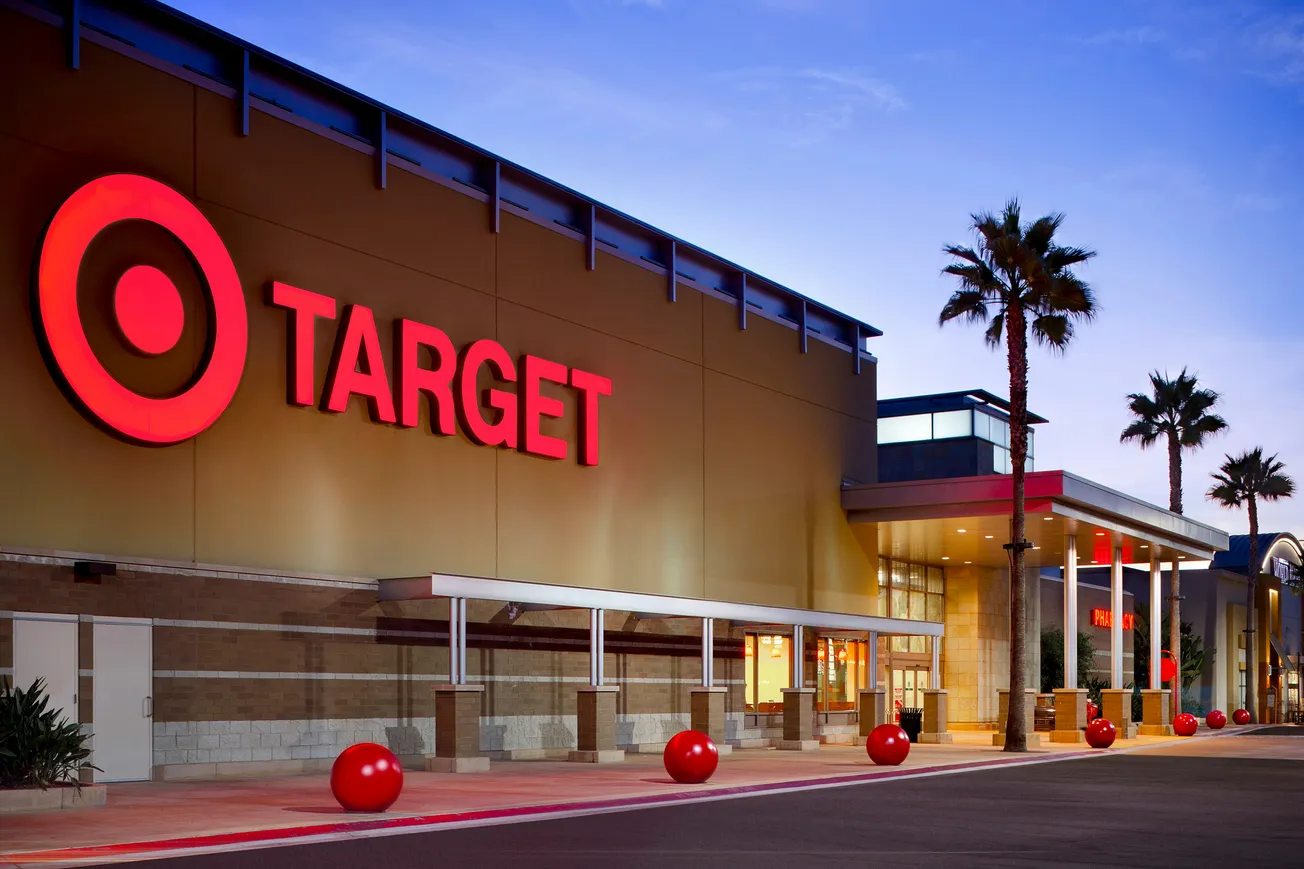 Storefront of a Target retail store at dusk. Red logo on the building, surrounded by palm trees and red bollards, under a blue sky.