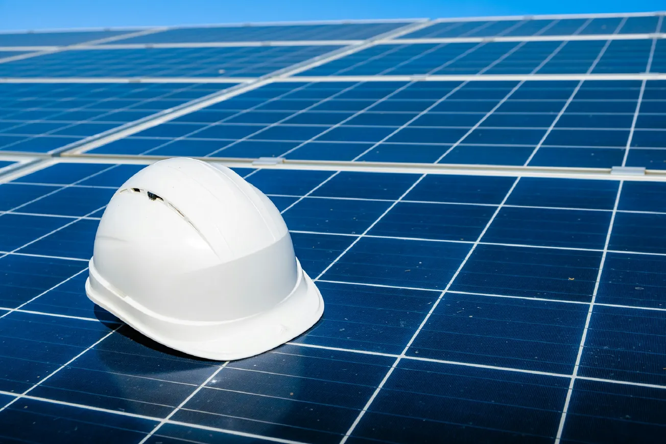 A white safety helmet rests on a field of shiny blue solar panels under a clear blue sky, symbolizing renewable energy and construction safety.