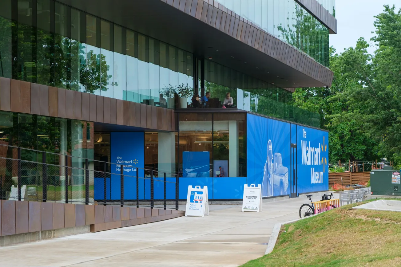 Glass-fronted building housing The Walmart Museum. Blue banners, entry signs, and modern facade. Trees in background, bicycle parked outside.